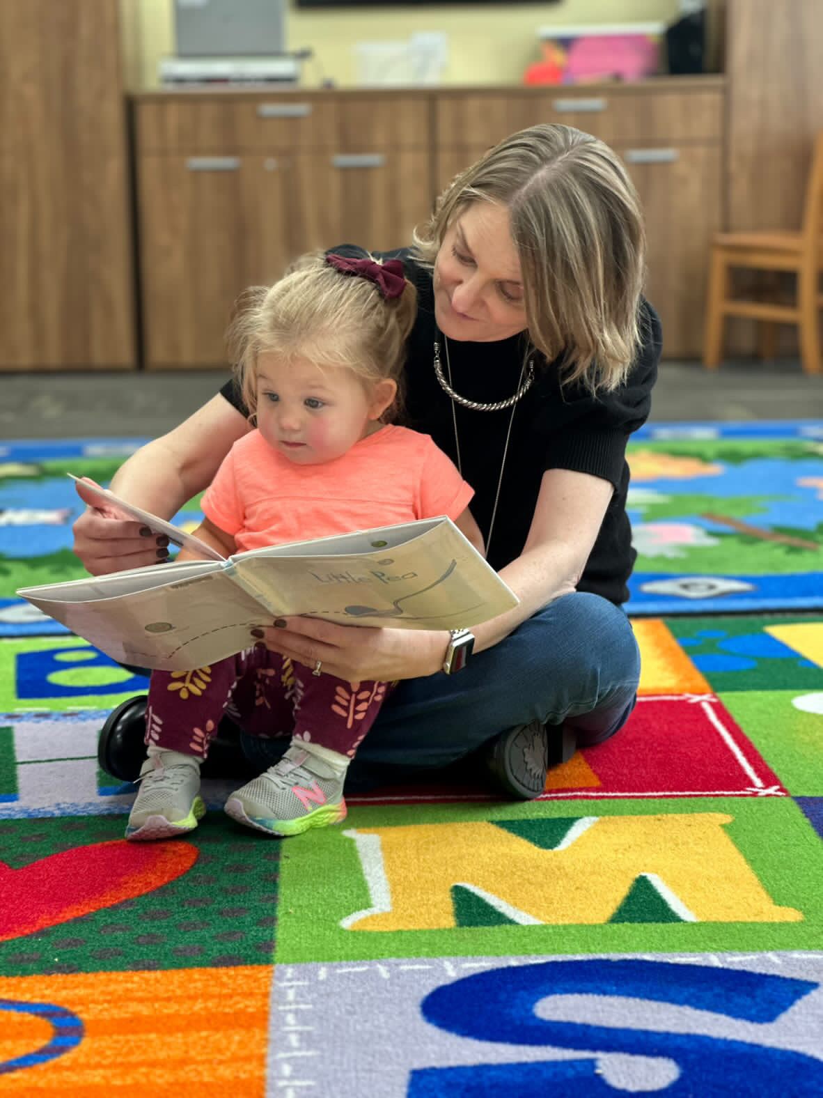 Woman in black shirt and jeans sitting on storytime rug with little girl in lap reading to her.