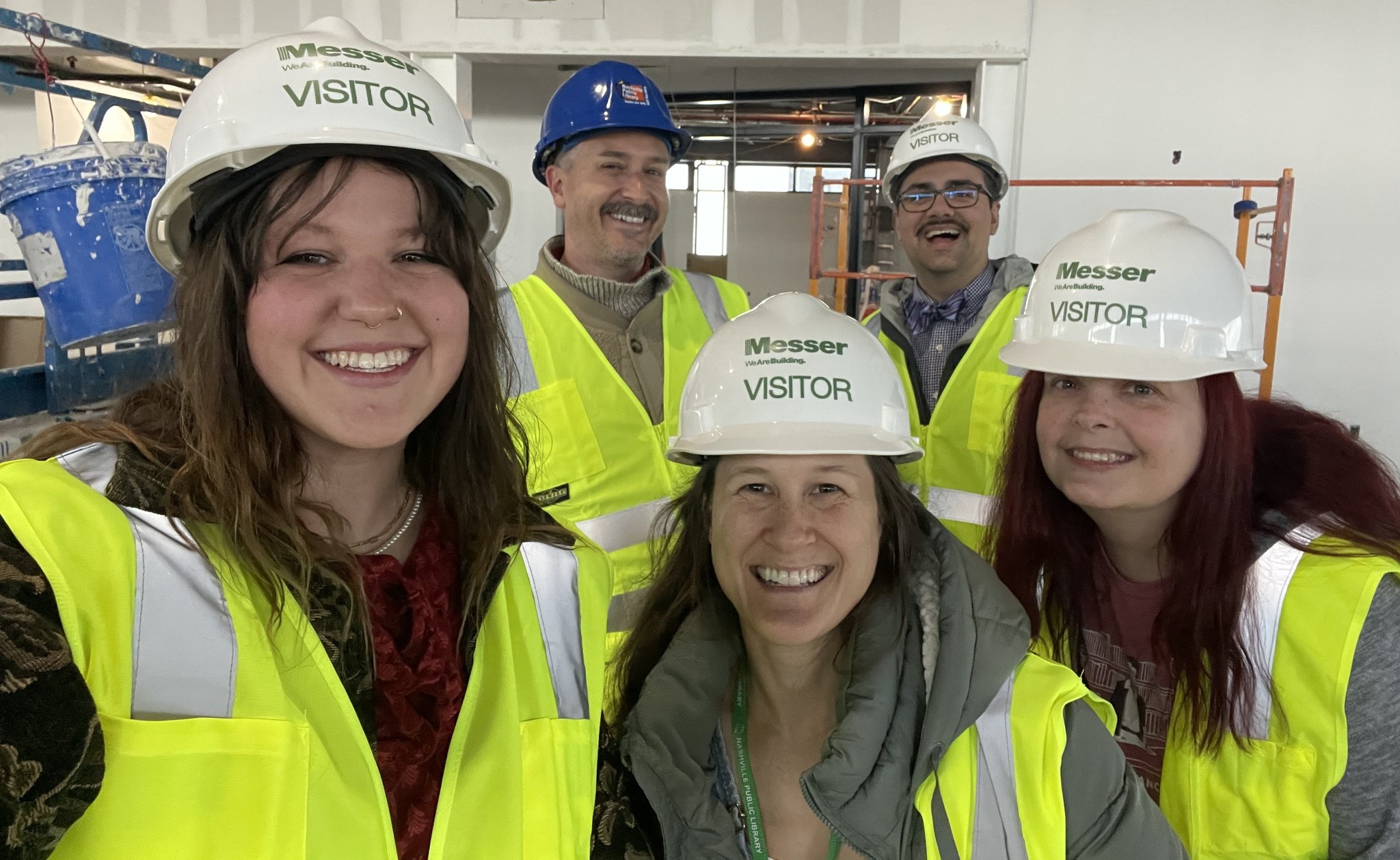 Five smiling people in an industrial setting wearing white and blue hard hats and high-visibility yellow safety vests while taking a group photo.
