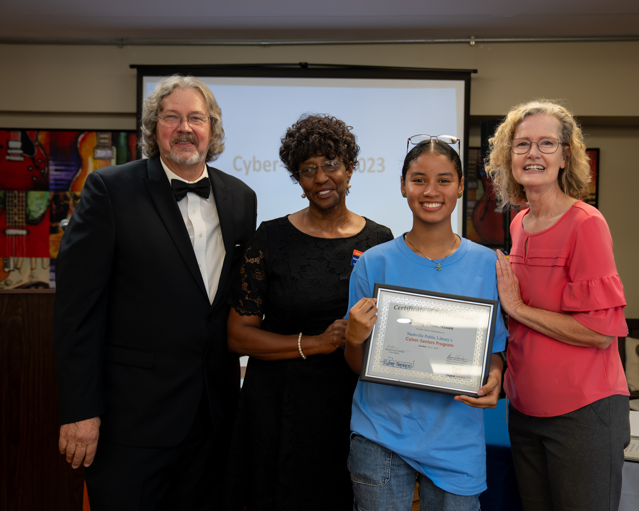 Four people standing together at an awards presentation, with one young woman in a blue shirt holding a framed certificate.