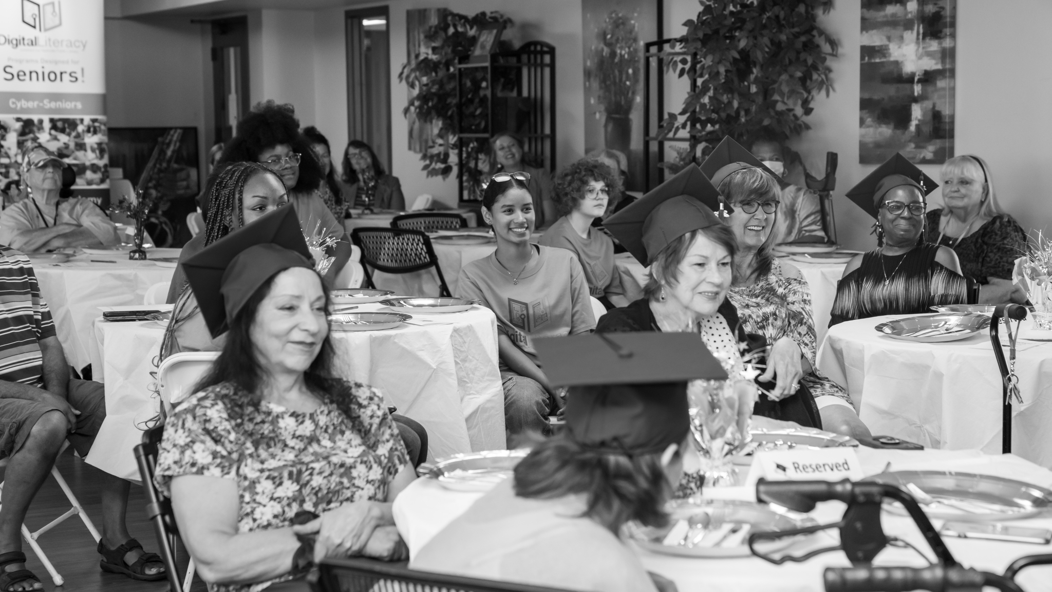 Black and white photo of a large group of people seated at tables in a room, with several women in the foreground smiling and looking toward a speaker.