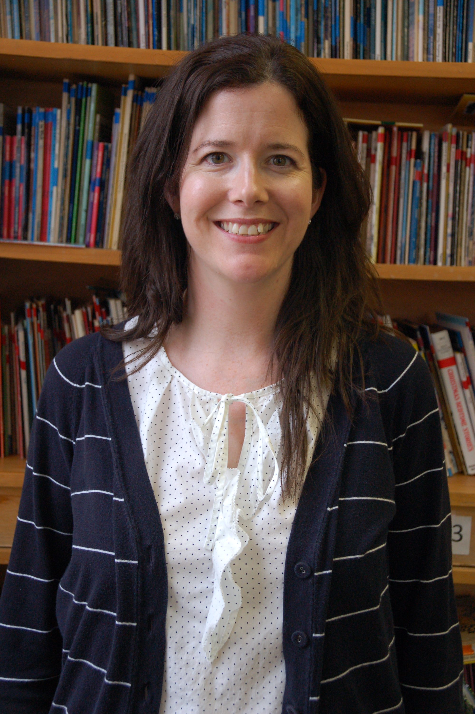 A smiling woman with long dark hair wearing a white blouse and a dark striped cardigan, standing in front of bookshelves filled with books.