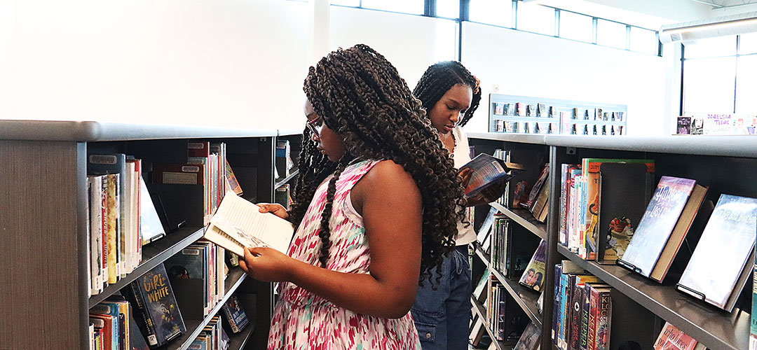 Two young black women stand between book shelves and read.