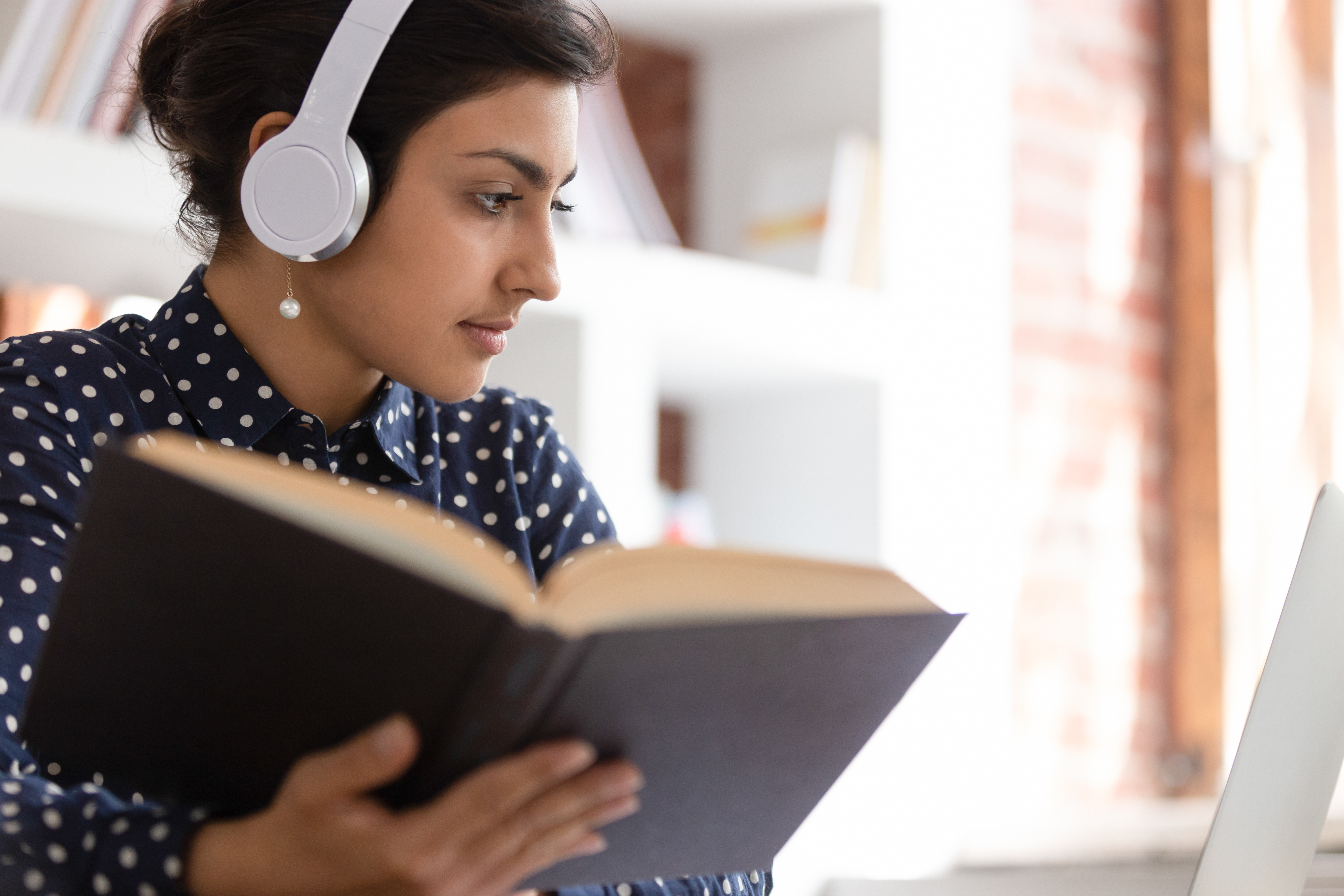 Woman reading while listening to headphones and looking at computer.