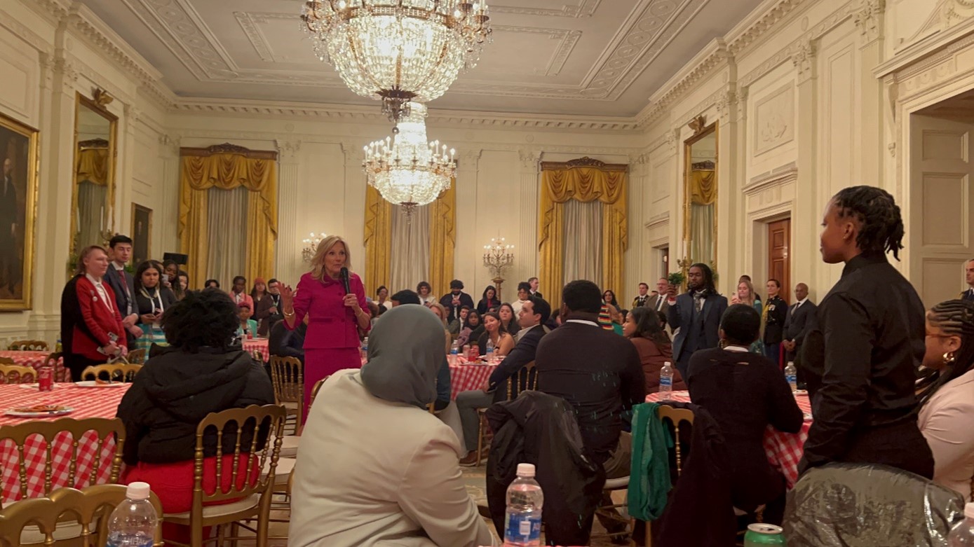 Group of adults and teens in a fancy room with gold curtains, chandeliers, gold chairs, and tables with red tablecloths. 