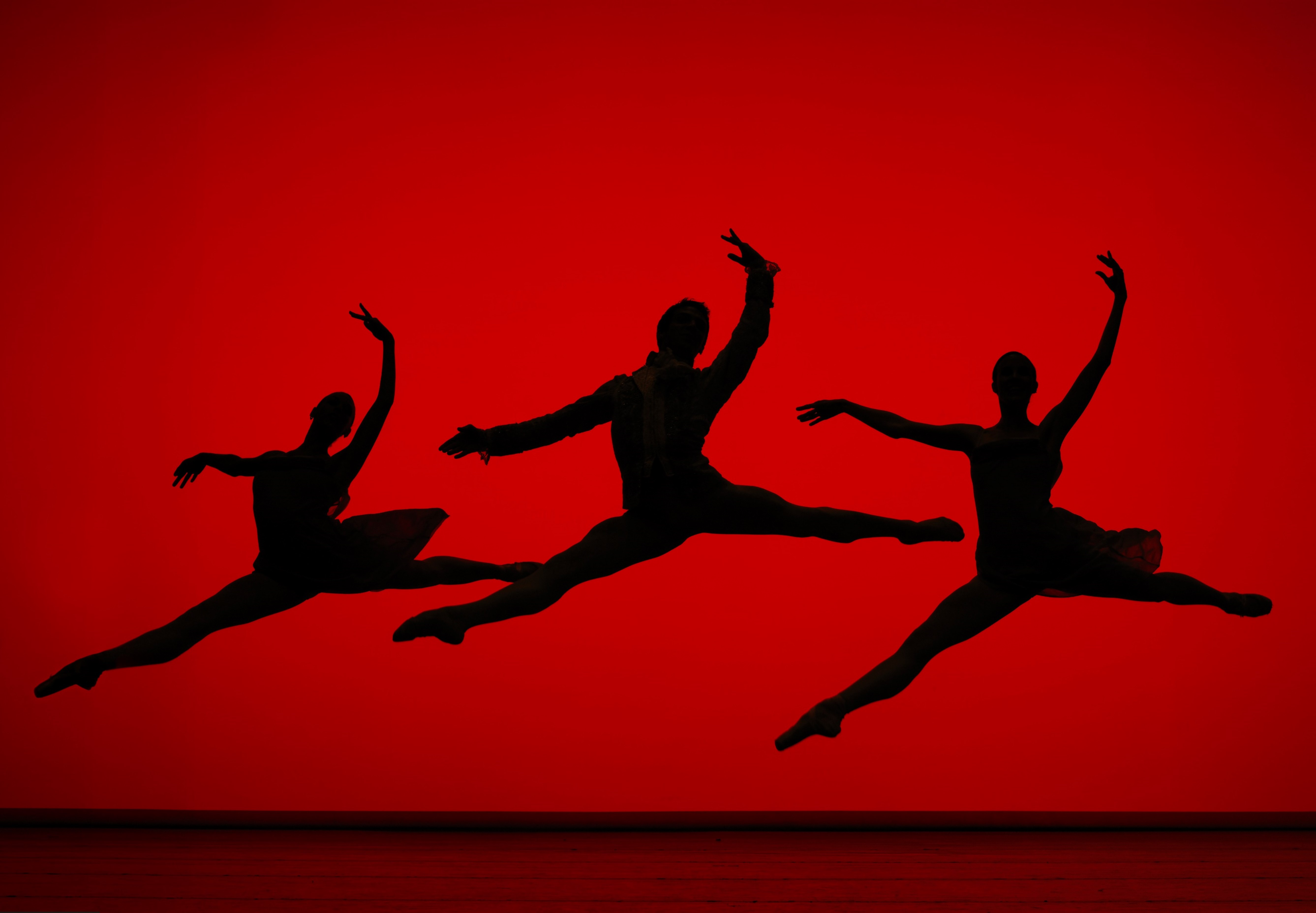 Silhouettes of three leaping ballet dancers against red backdrop.
