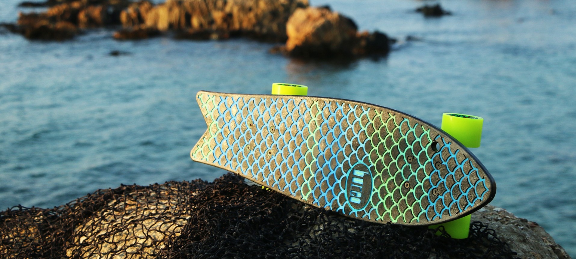 Fish-shaped skateboard with fish scales on its side on rock by ocean.