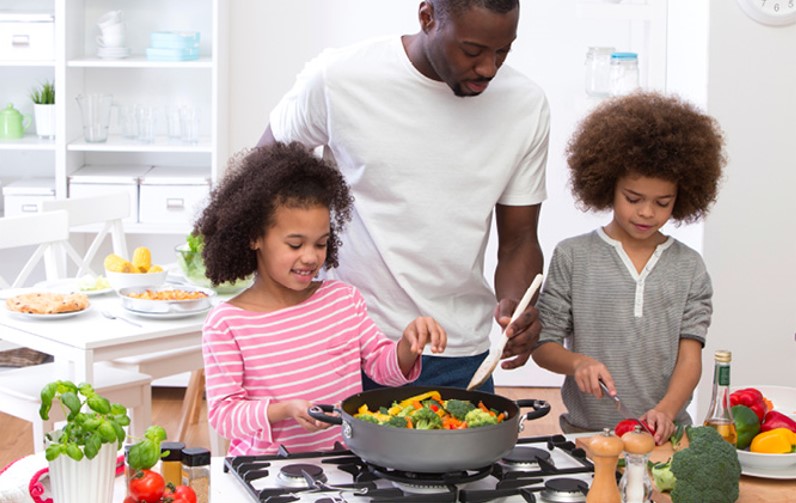 A man and two young children cook together in a bright kitchen. The daughter stirs a pan of green vegetables while the father guides her.