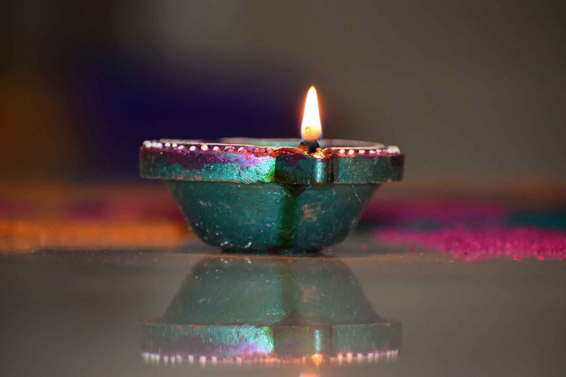 A single lit diya lamp with a bright flame rests on a reflective surface against a dark, colorful background, symbolizing light and celebration.