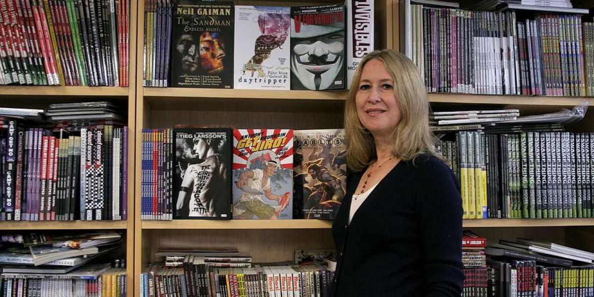 A blonde woman in a black blazer stands smiling in front of wooden bookshelves filled with various books in a library or bookstore.