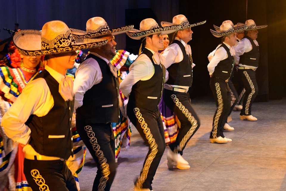 A line of performers in traditional black and white outfits and hats perform a synchronized dance on a stage with dramatic lighting.