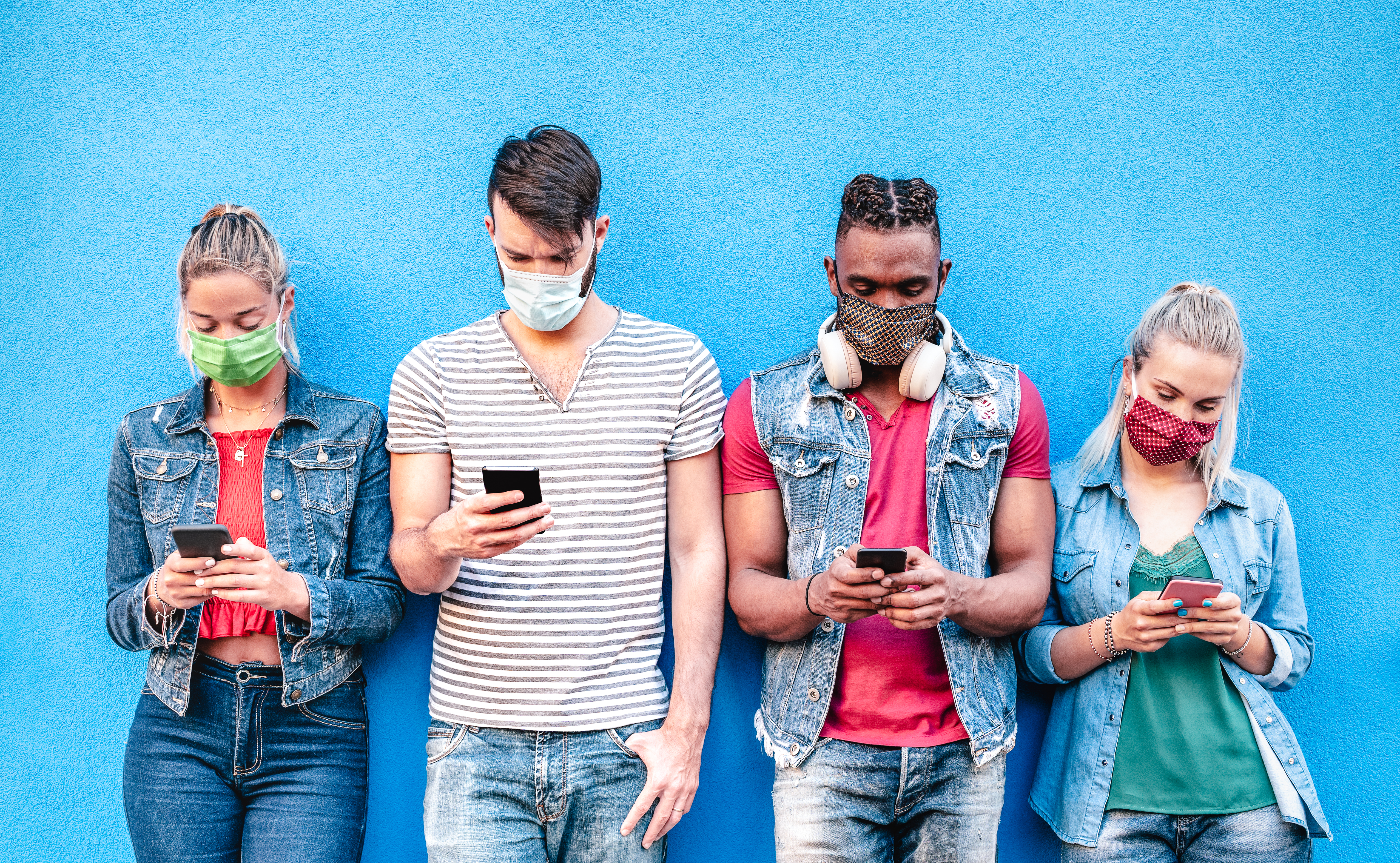 Four diverse young adults wearing protective face masks stand against a bright blue wall, each looking down and focused on their smartphones.