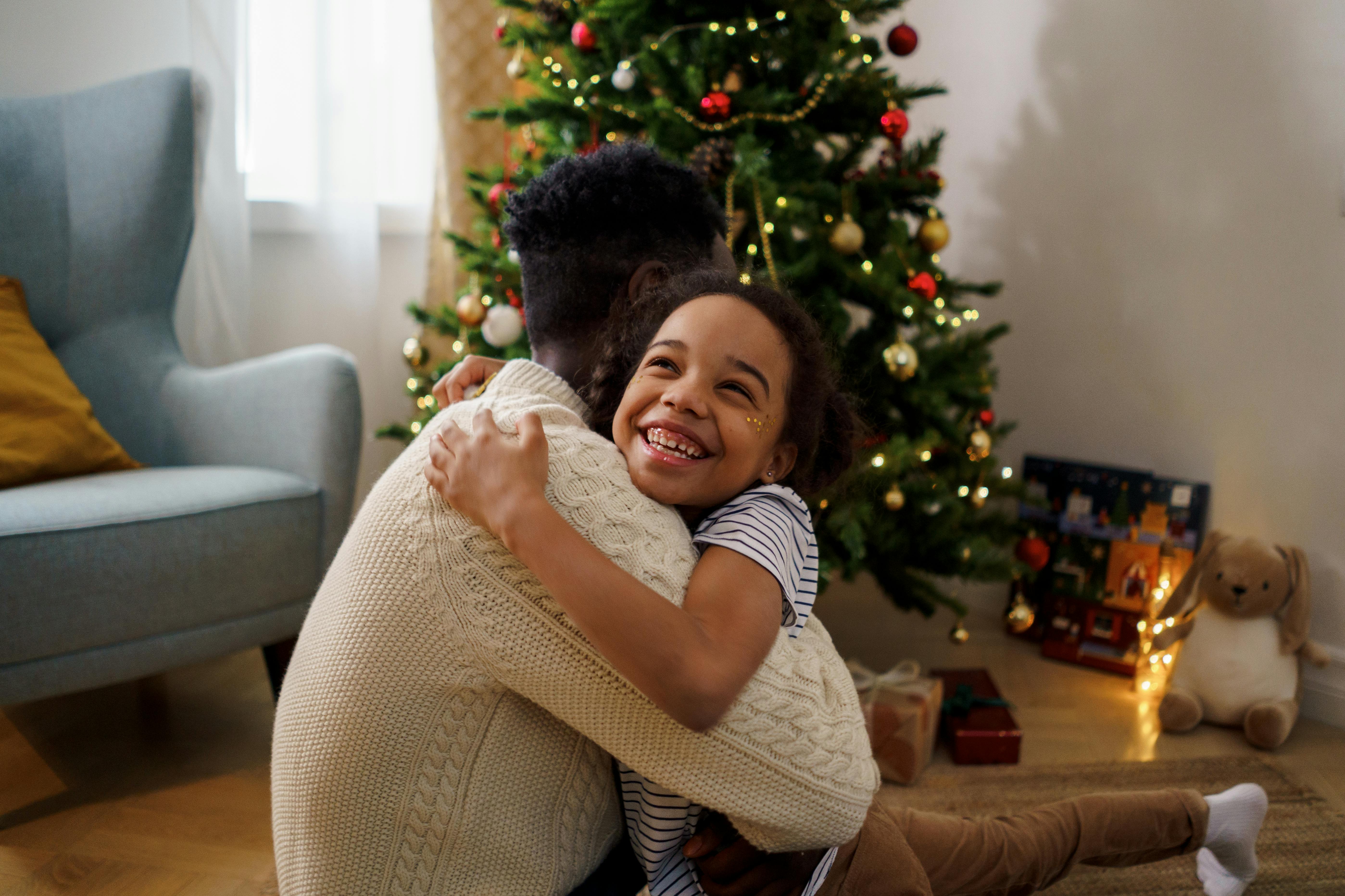 A young girl with a happy face hugs a man in a cream cable-knit sweater. They are sitting on the floor in a cozy living room in front of a decorated Christmas tree glowing with lights.