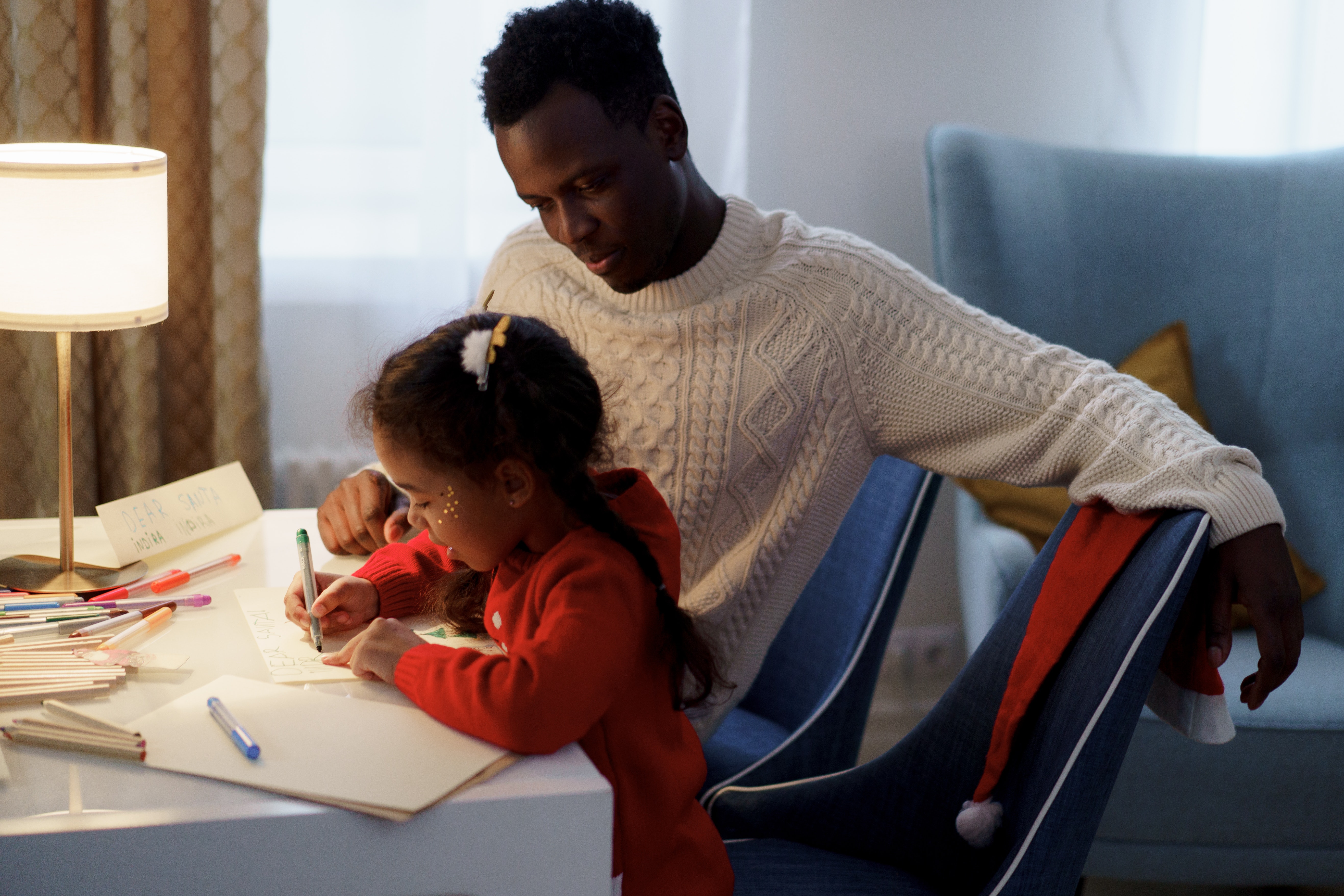 A young girl with glitter on her face sits at a table next to a man in a cream cable knit sweater. The girl is writing on paper with a green marker. 