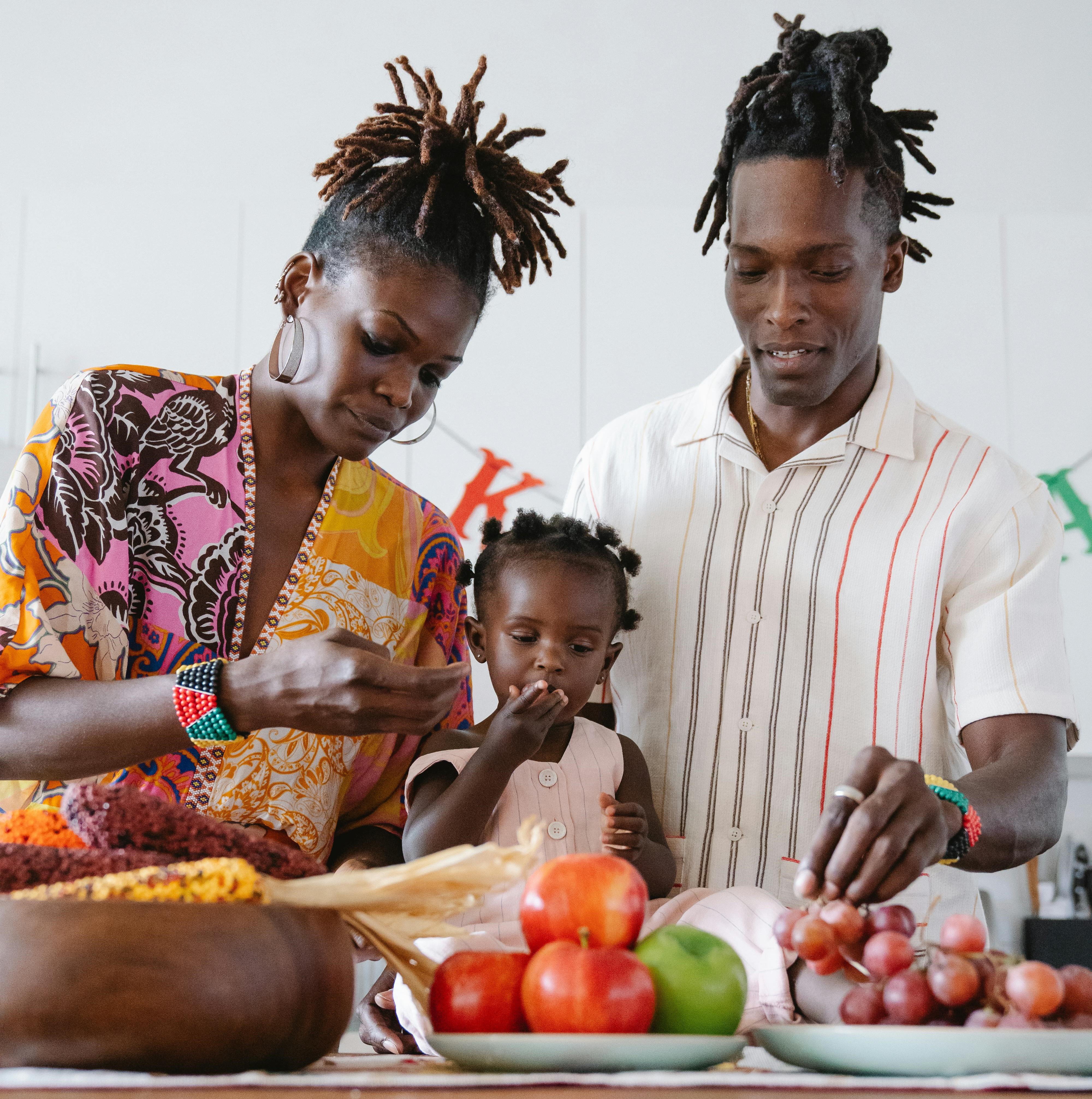 A young girl stands at a kitchen island and eats a grape. A man and a woman stand on either side of her. There are apples and grapes and corn in the foreground.