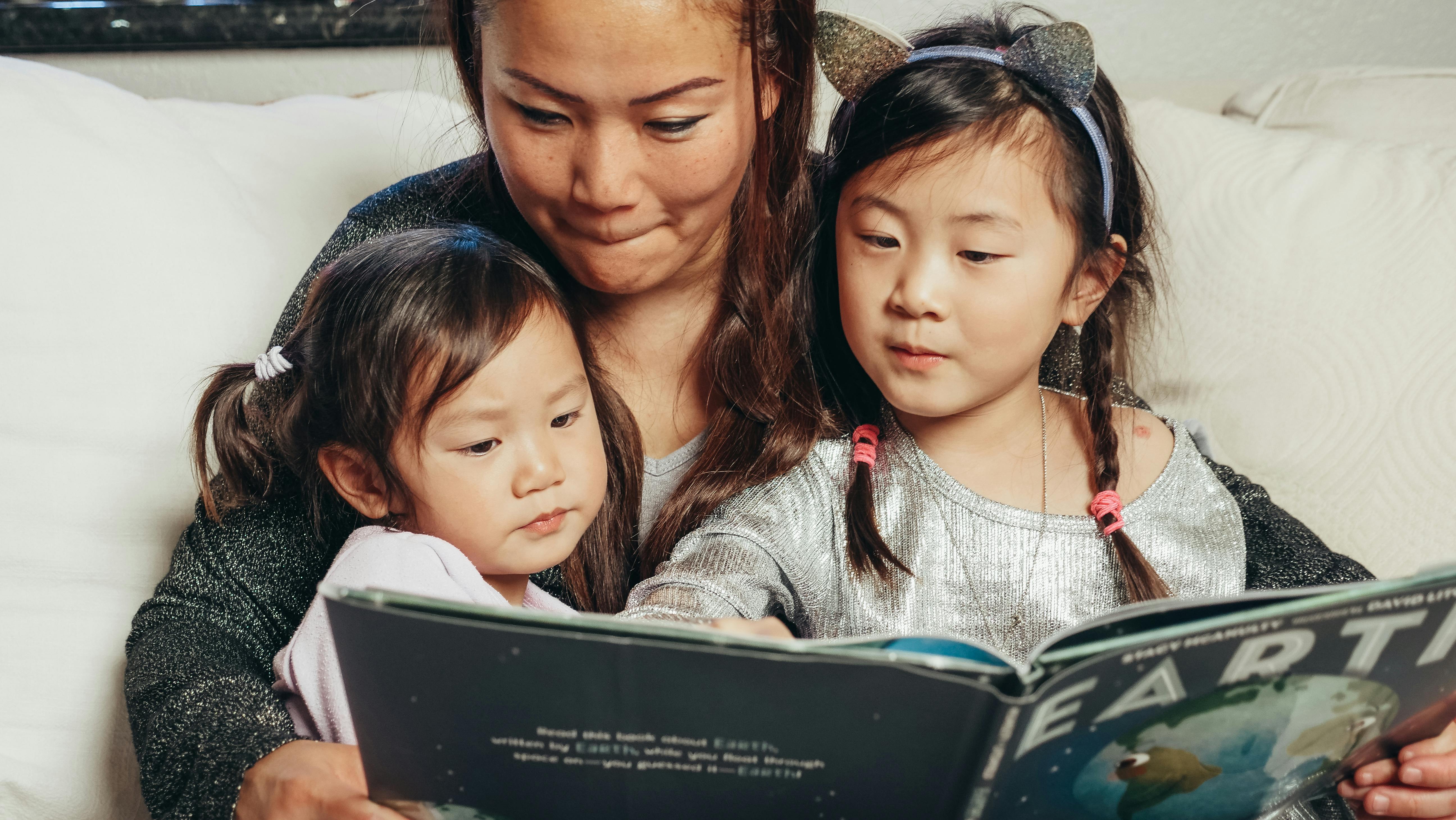 A woman sits with two little girls in her lap. The girls hold an open book called Earth.
