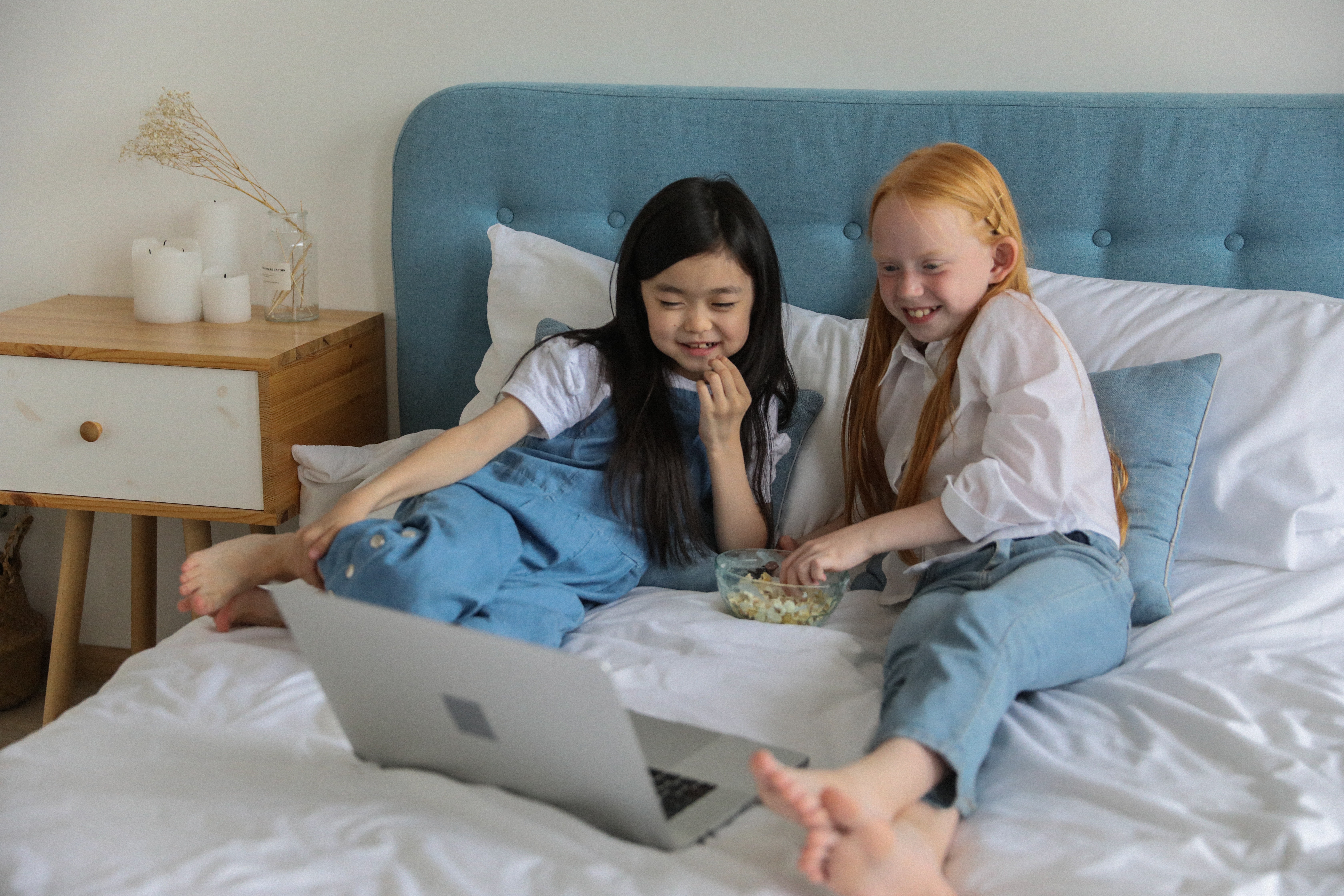 Two young girls lay on a blue bed eating popcorn from a clear glass bowl while watching something on an open laptop.