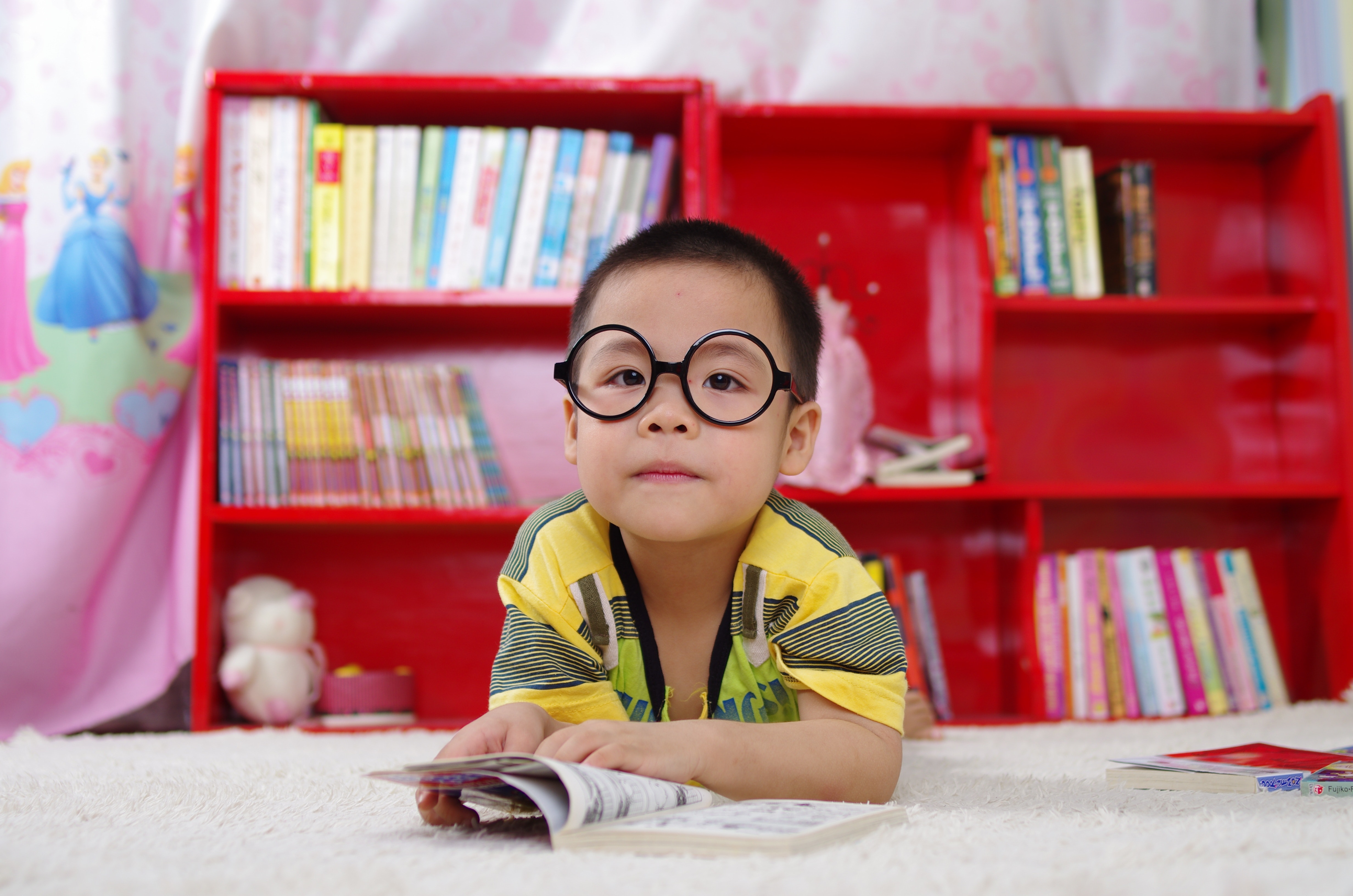 A young boy wearing dark glasses lays on a cream color carpet and reads a book. There are red bookshelves in the background.
