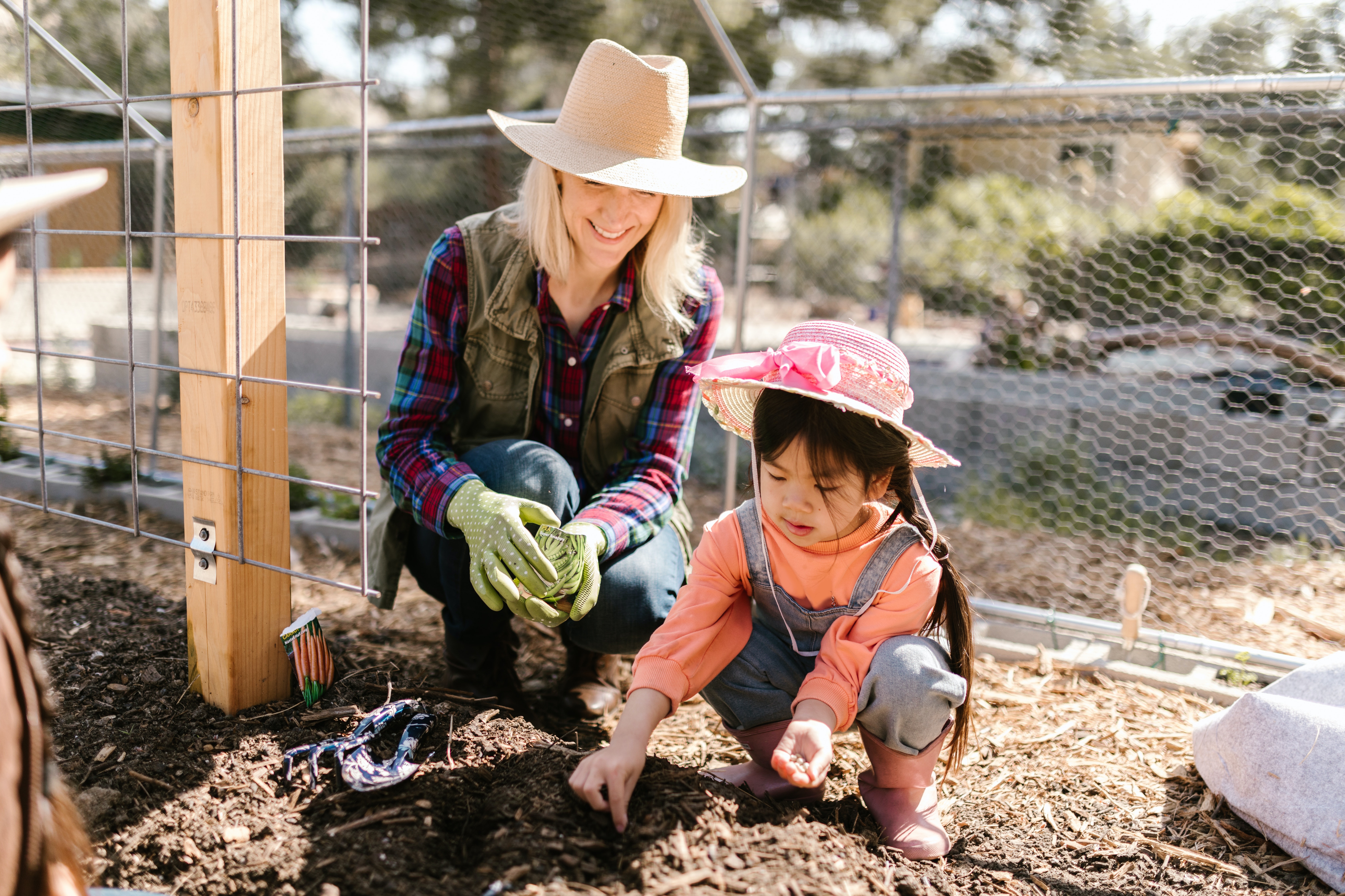 A blonde woman in a straw hit kneels next to a little girl with a pink straw hat while they plant seeds in the dirt.