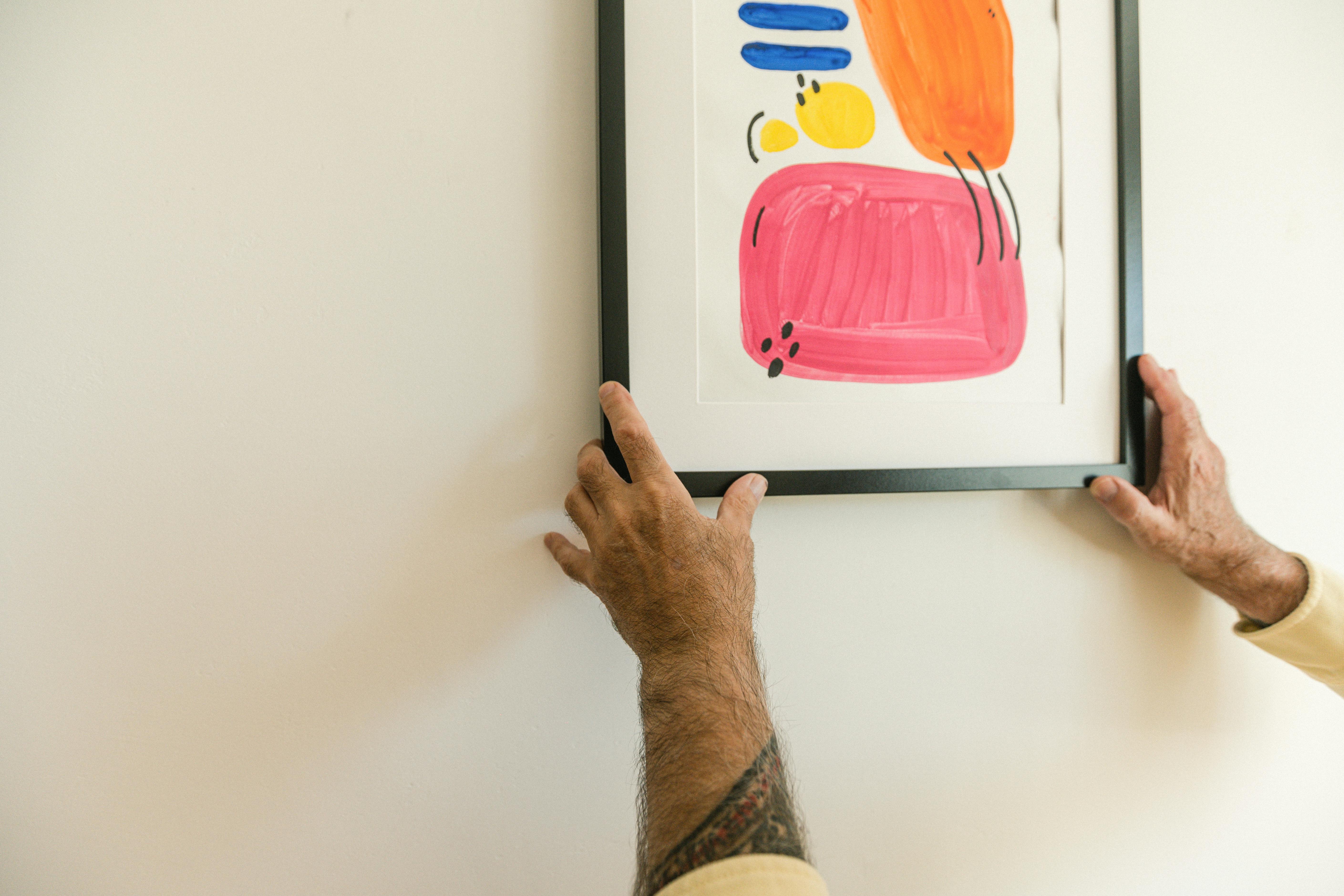 Two arms hang a colorful picture on the wall. The picture is an abstract painting with pink, orange, yellow, and blue dots.