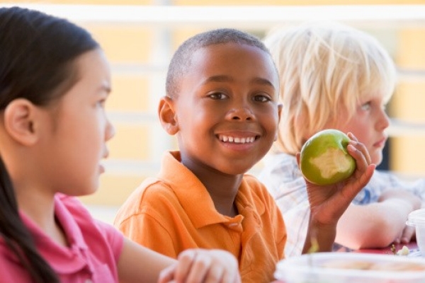 Three children sit together and the boy in the middle eats a green apple