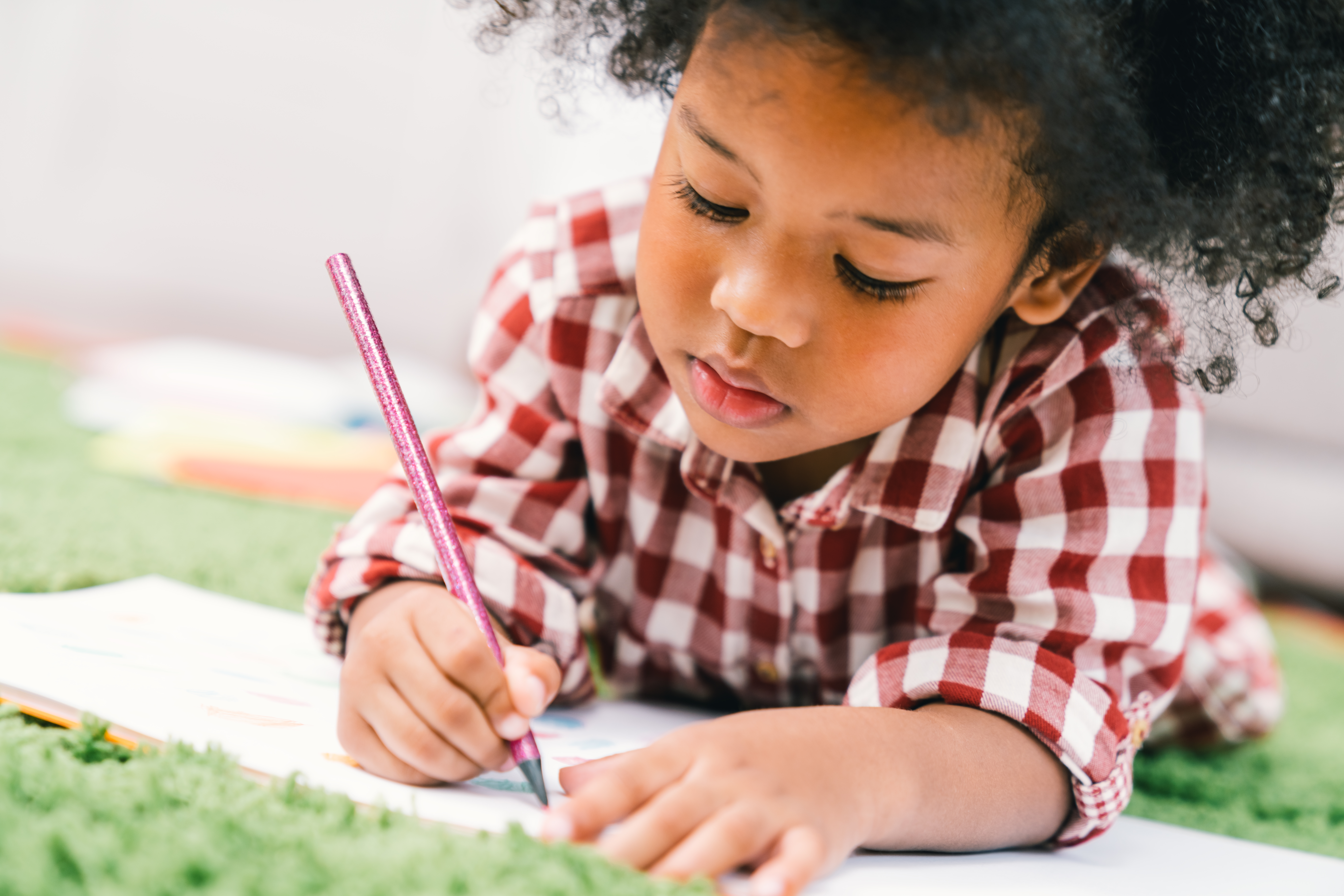 Preschool age child laying on a blanket drawing on a paper with a pen.