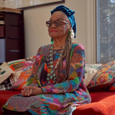 Image of artist Faith Ringgold. She is a slim older African American woman, seated on a red couch. She wears a printed dress, a blue head wrap, and glasses. 