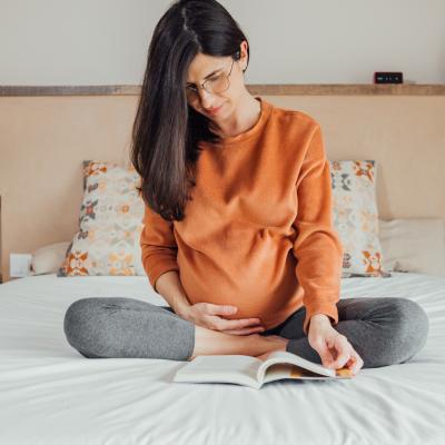 Pregnant woman reading book while sitting on a bed.