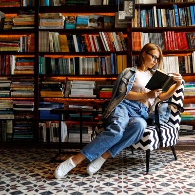 Girl in a black and white zig zag chair sits reading in front a large bookshelf.