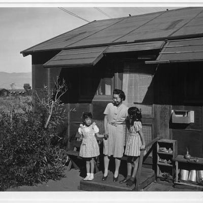 Photograph by Ansel Adams, 1943. Black and white photograph shows Mrs. Yaeko Nakamura and her two children, Joyce Yuki (right) and Louise Tami (left), standing on the step at the entrance of a wooden dwelling, Manzanar Relocation Center. 
