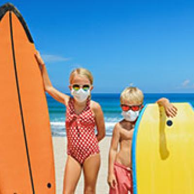 Two girls with surfboards on the beach wearing masks over their mouth and nose.