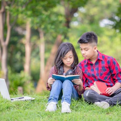 Young boy and girl sit in a park reading and playing on a computer