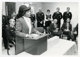 woman stands at a lecturne. Men and women in civil law enforcement uniforms look on.