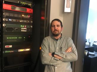 Slightly smiling man standing with arms folded at chest in front of electronic console wall.