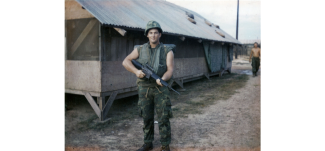 White man in green military fatigues holding rifle in front of tin roof building.