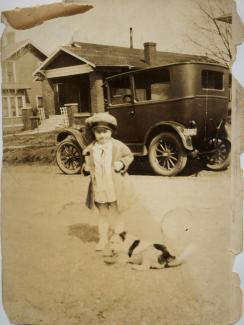 Black and white historical photo of a young boy and a dog in front of a car.