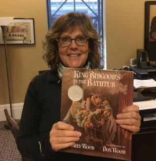 A smiling woman with glasses and curly hair holds up a children's picture book titled "Finding Winnie" in an office setting.
