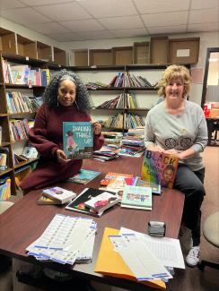 Two women sitting on a desk in a room filled with books, each holding a picture book.