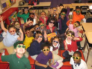 Smiling young students wearing sunglasses, sitting on a carpet around a woman holding a book.