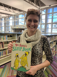 A smiling woman in a library holds up Dr. Seuss's Sleep Book. She wears a patterned shirt, a cream knit scarf, and glasses perched on her head.