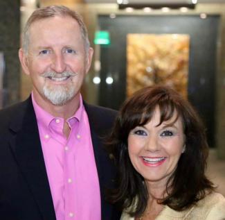 A smiling man with a grey beard in a pink shirt and dark blazer stands next to a woman with brown hair and bangs in an indoor setting.