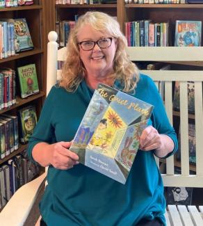 Woman, sitting on a bench, holds childrens book. Is seated in a library.