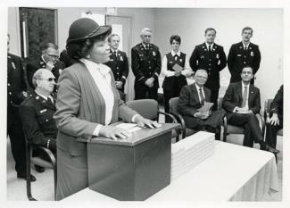 Black woman, Thelma Harper, stands at a podium in a room of white men and women