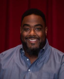 Smiling bearded Black man in blue shirt standing in front of red velvet curtain.